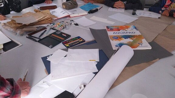 Une table avec divers matériaux de dessin, y compris des carnets, des papiers, des crayons et de la peinture, entourée de participants à un atelier d'illustration.